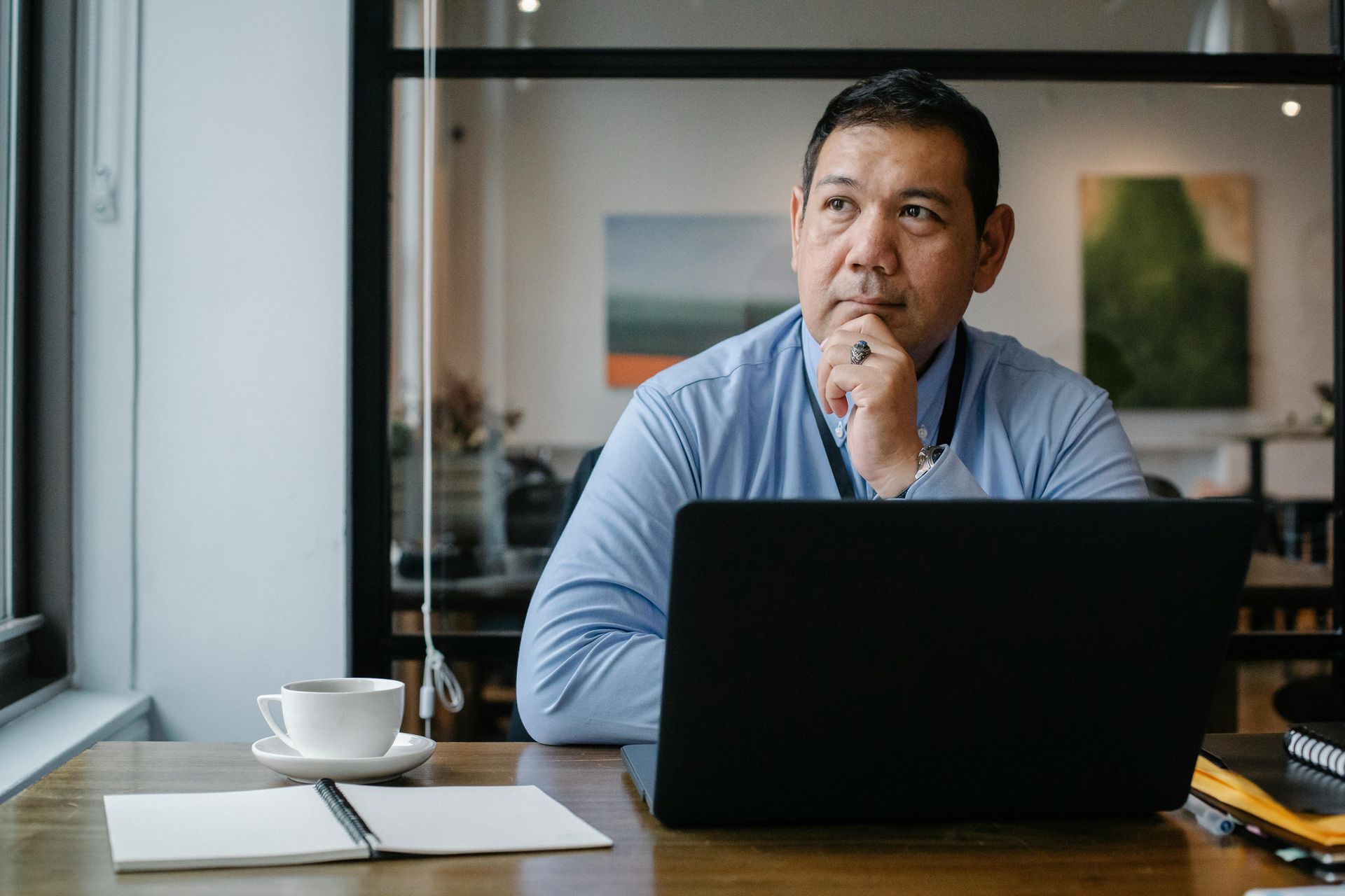 Man at desk, laptop open, looking thoughtful, hand on chin. Cup of coffee, notebook visible.