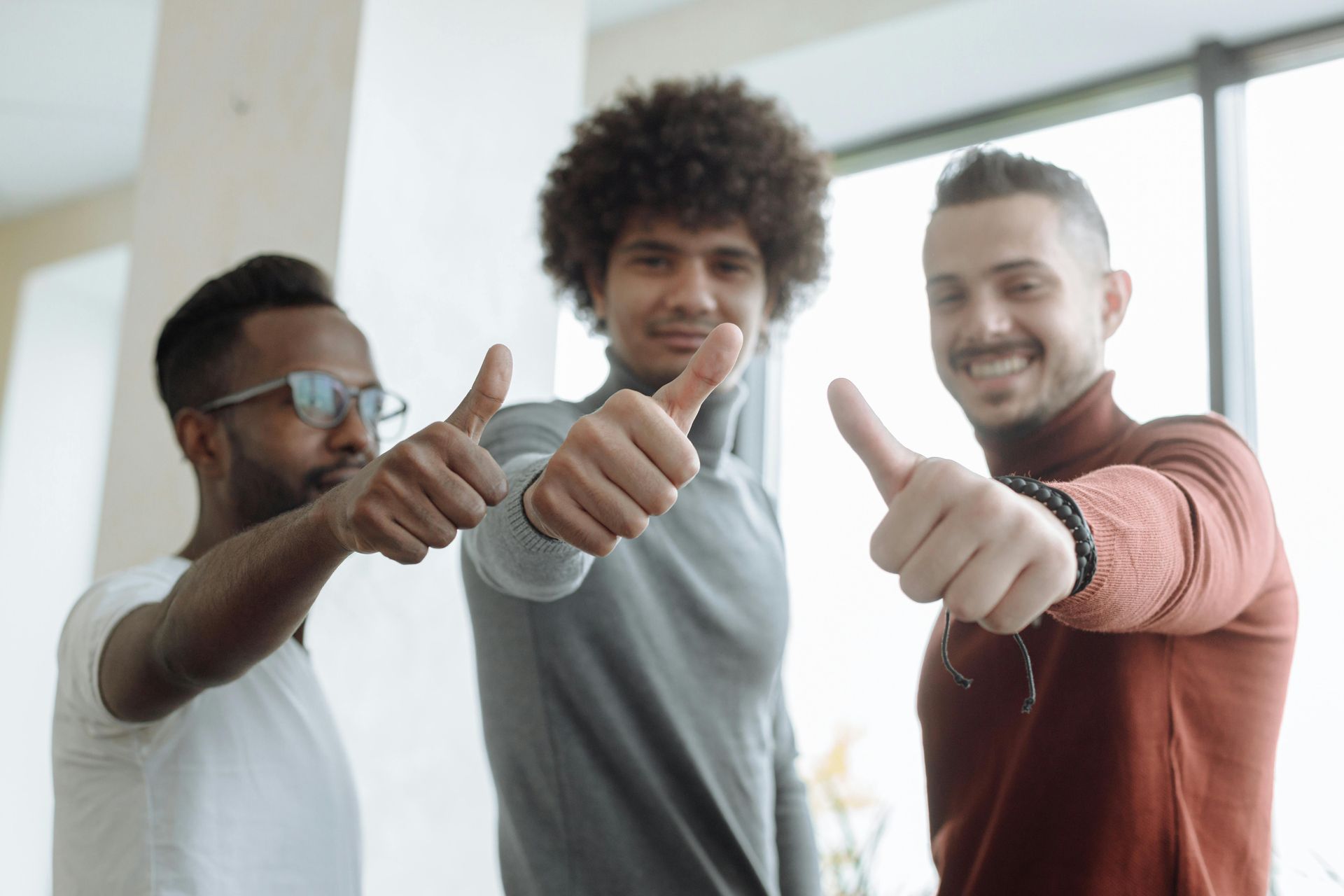 Three people giving thumbs-up gestures, smiling. Standing near a window.