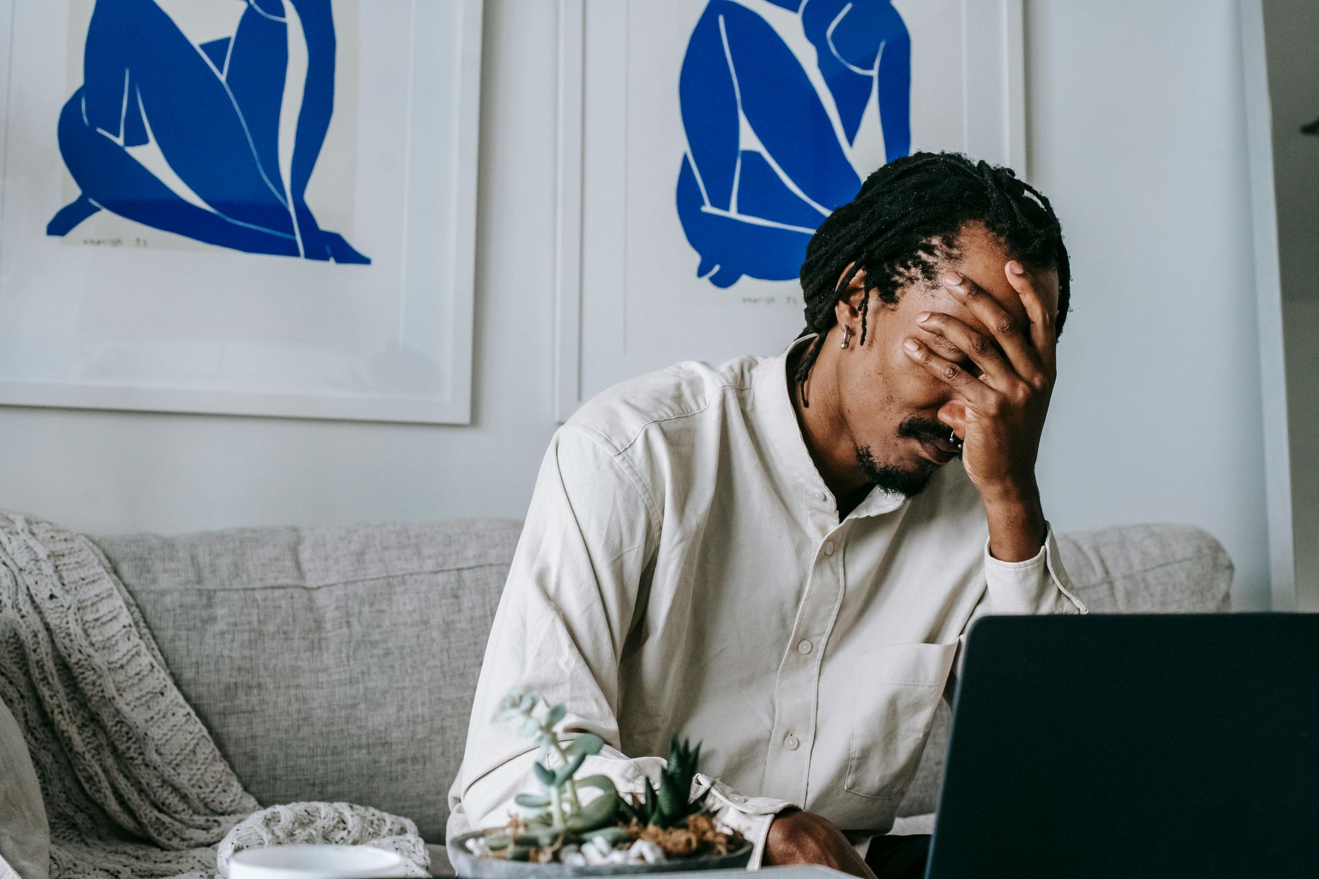 Man sitting on couch, head in hand, laptop in front; appears stressed, two blue art pieces in background.