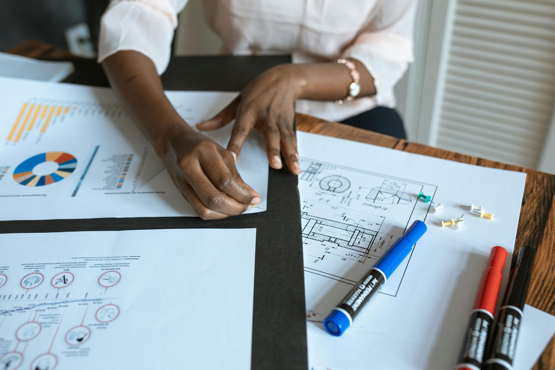 Hands work with markers and documents featuring charts and diagrams on a wooden desk.