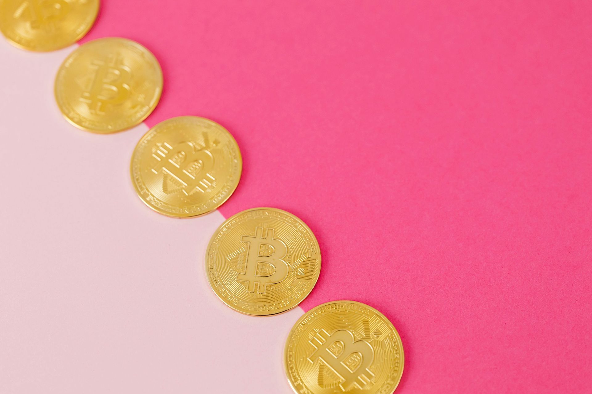 Five gold-colored Bitcoin tokens lined up diagonally across a split background of light pink and deep pink.
