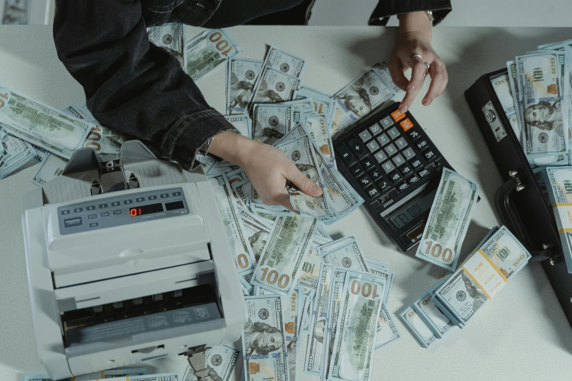 A person using a calculator and money counter surrounded by stacks of US hundred-dollar bills on a table.