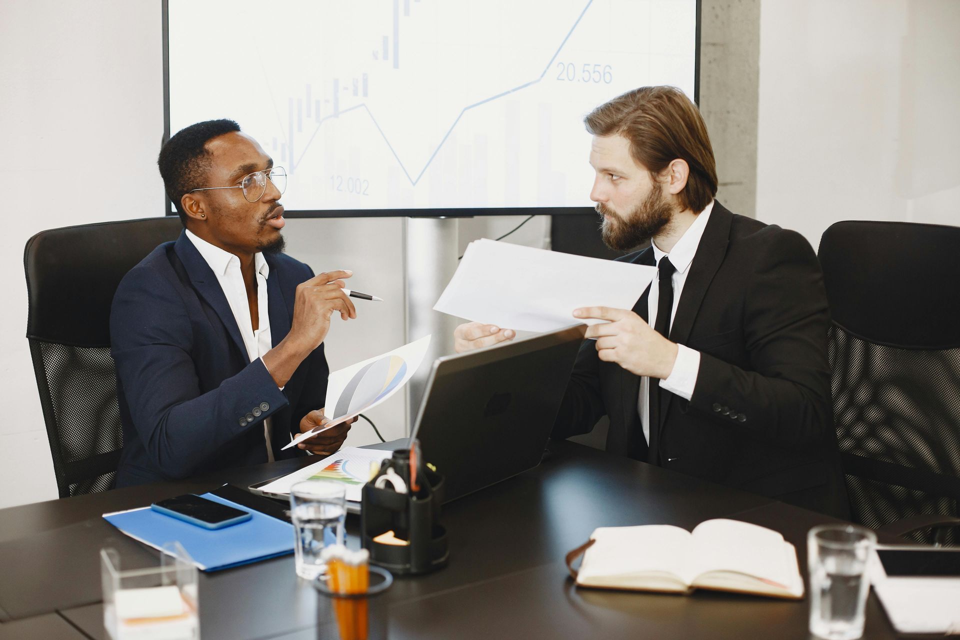 Two business professionals in suits discuss documents at a table with a laptop, with a graph displayed on a background screen.