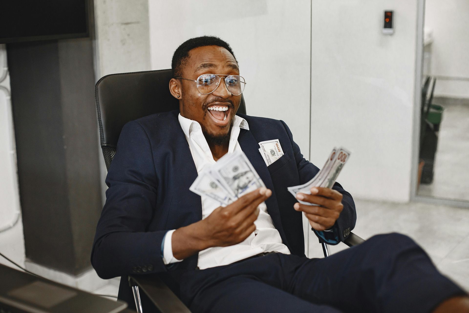 A person in a suit and glasses laughs while holding stacks of US dollar bills in an office setting.