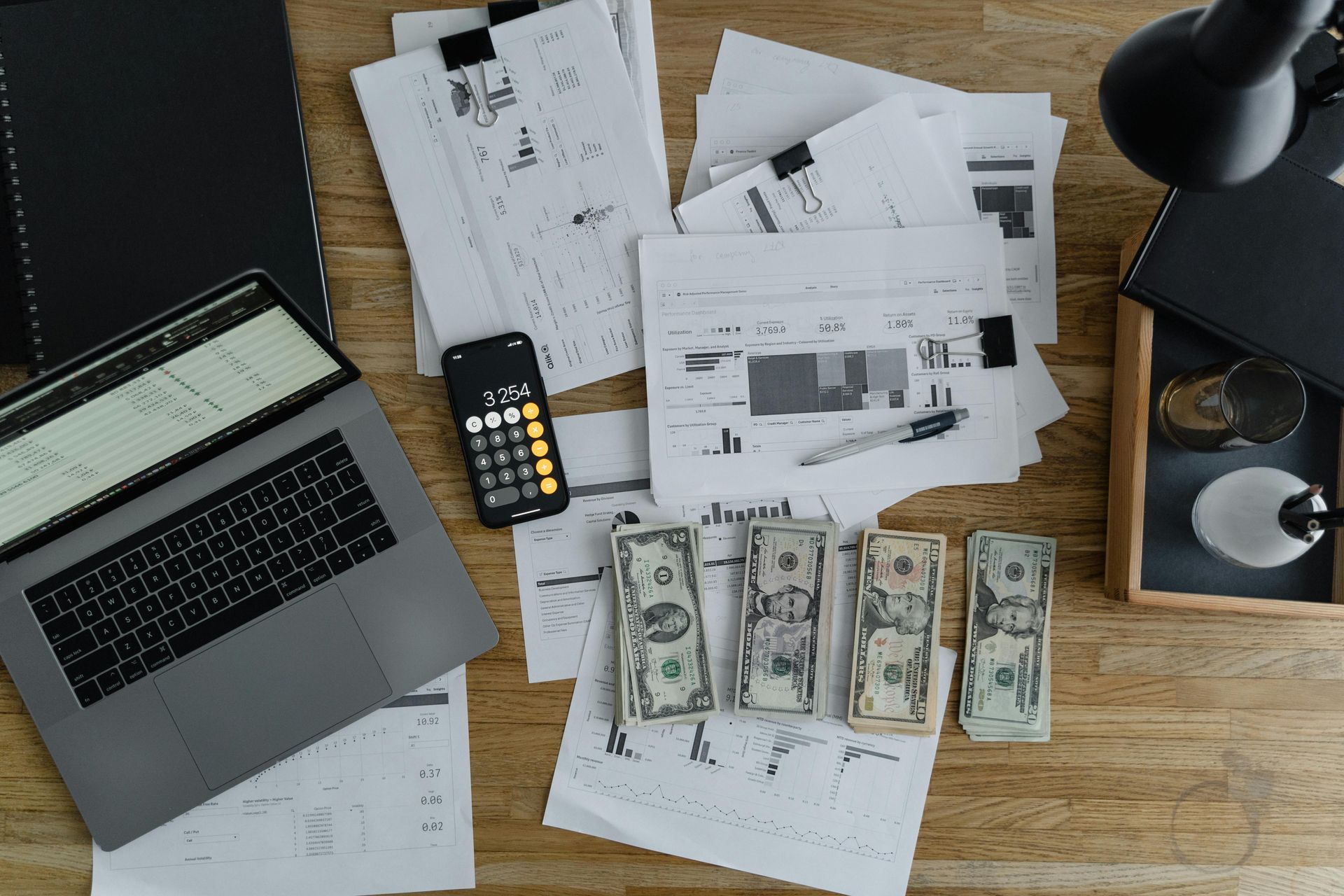 A top-down view of a wooden desk featuring a laptop, calculator, scattered paperwork, and four stacks of paper money.