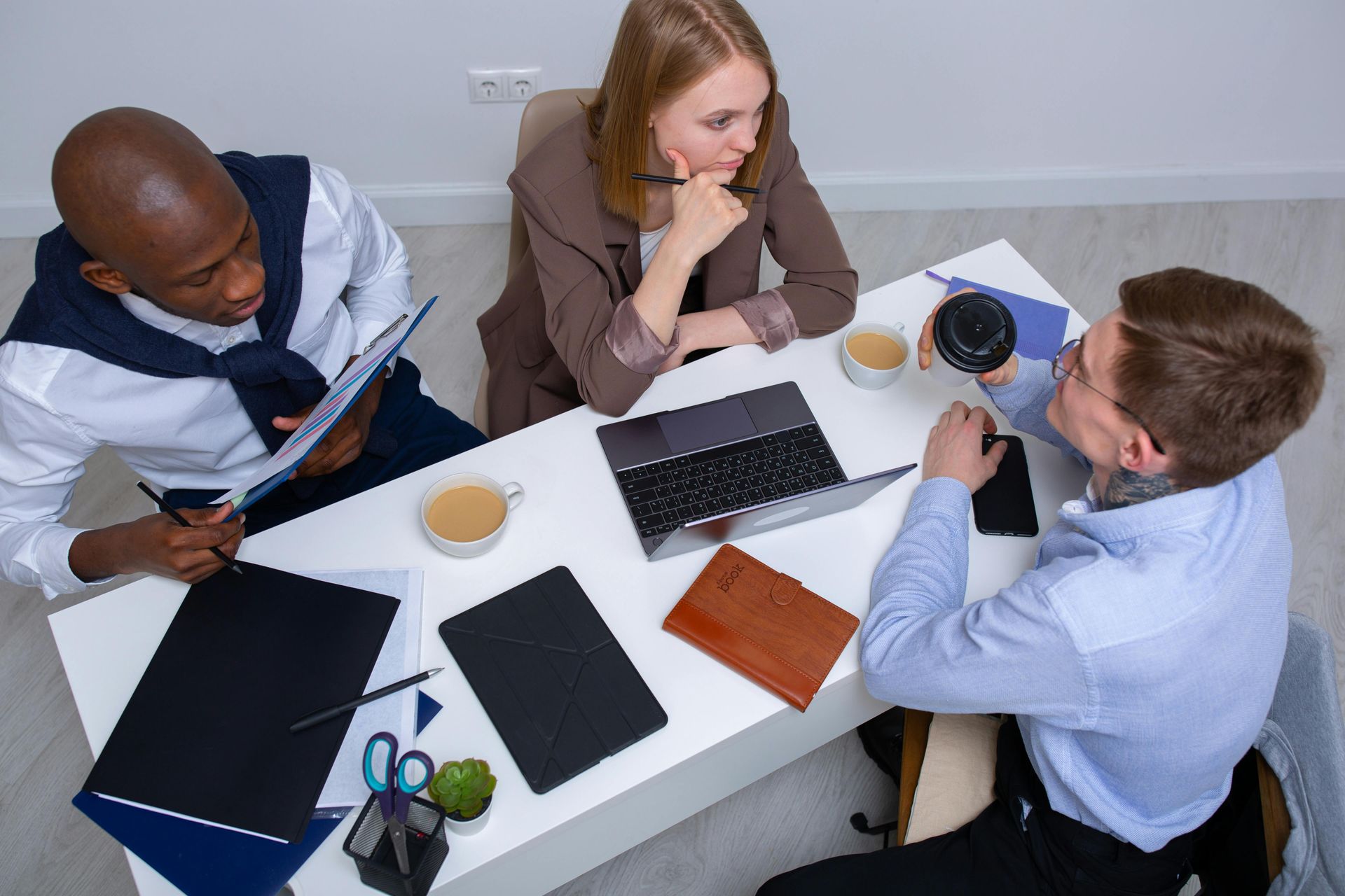 Three people sit at a white table during a business meeting, reviewing documents and discussing work with laptops.