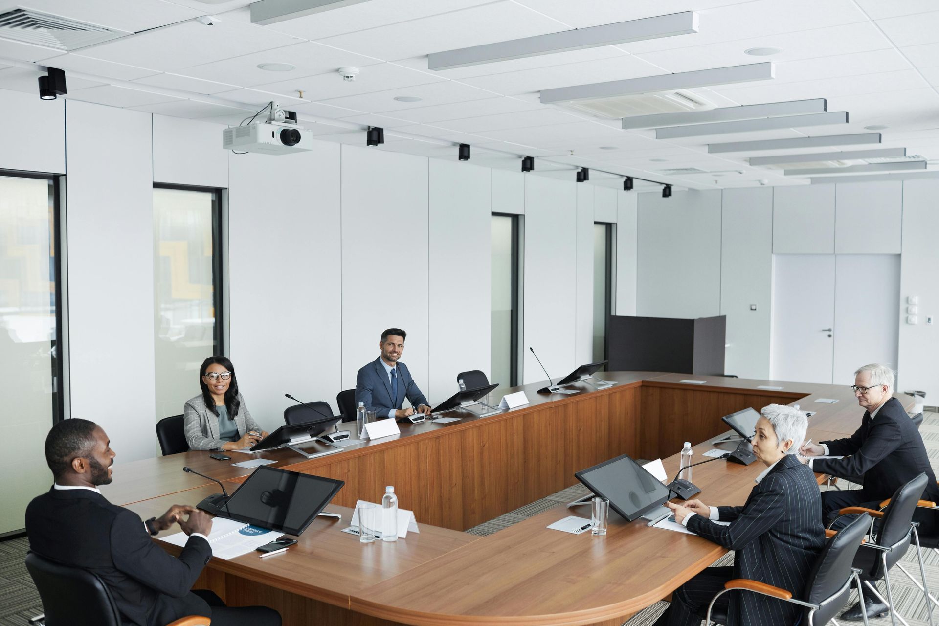 A diverse group of professionals in suits sitting around a U-shaped wooden table in a bright, modern conference room.