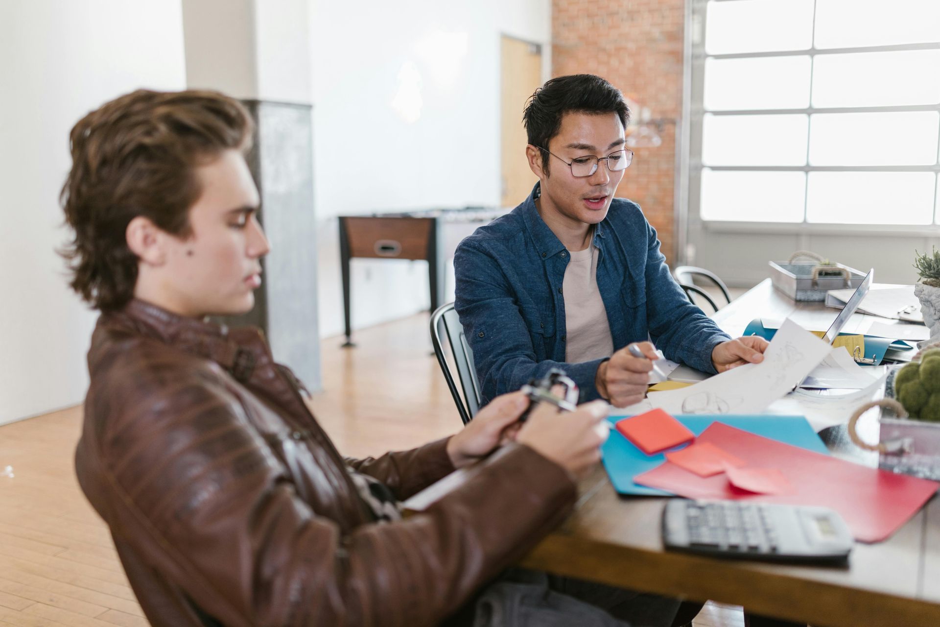 Two people sit at a table with papers, pens, and colorful sticky notes, collaborating in a bright, modern office space.