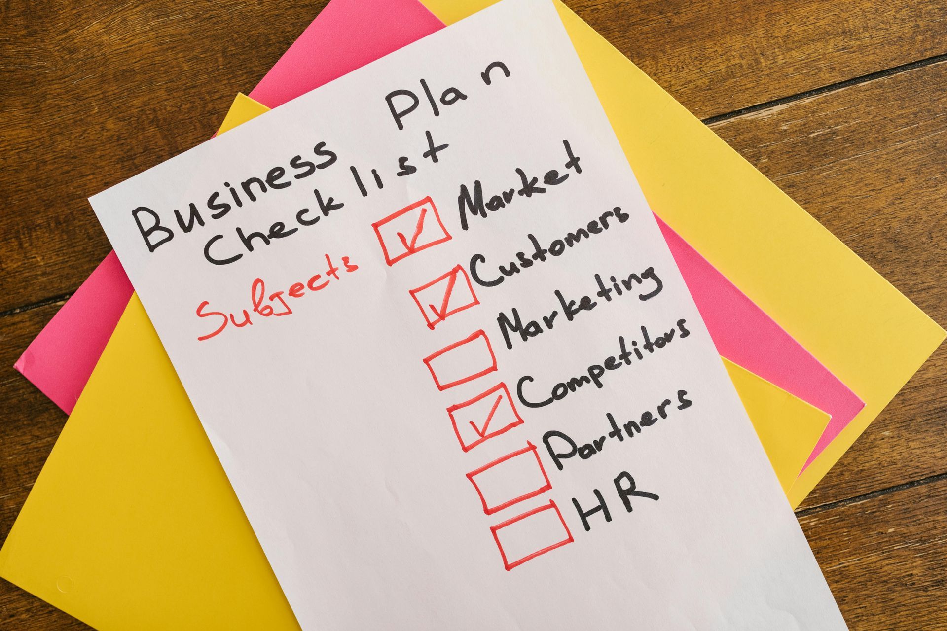 Business plan checklist with checked boxes for Market, Customers, and Competitors. Paper on colorful sheets.