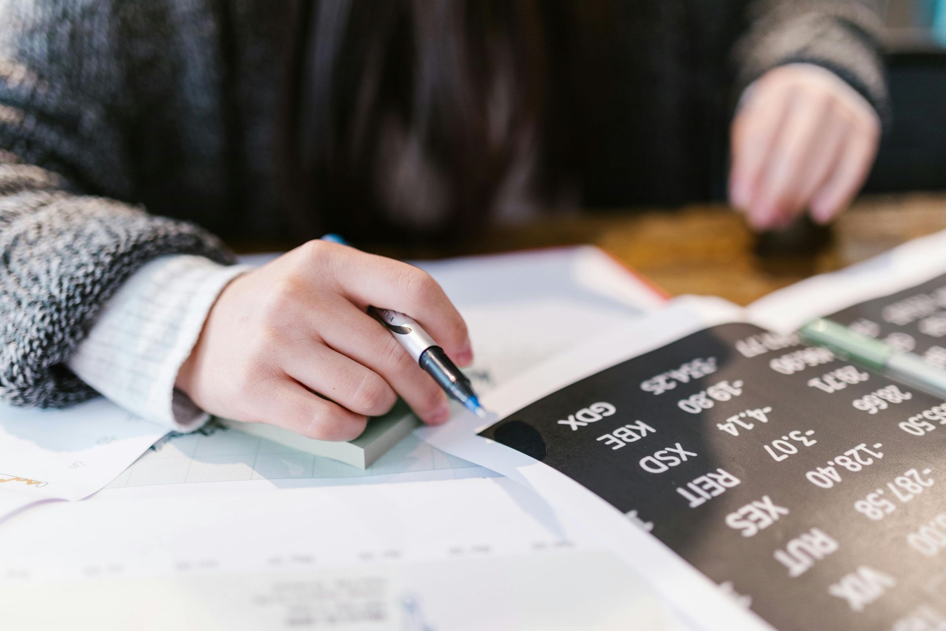 Hand writing on papers with a pen, next to a notebook with stock market information.