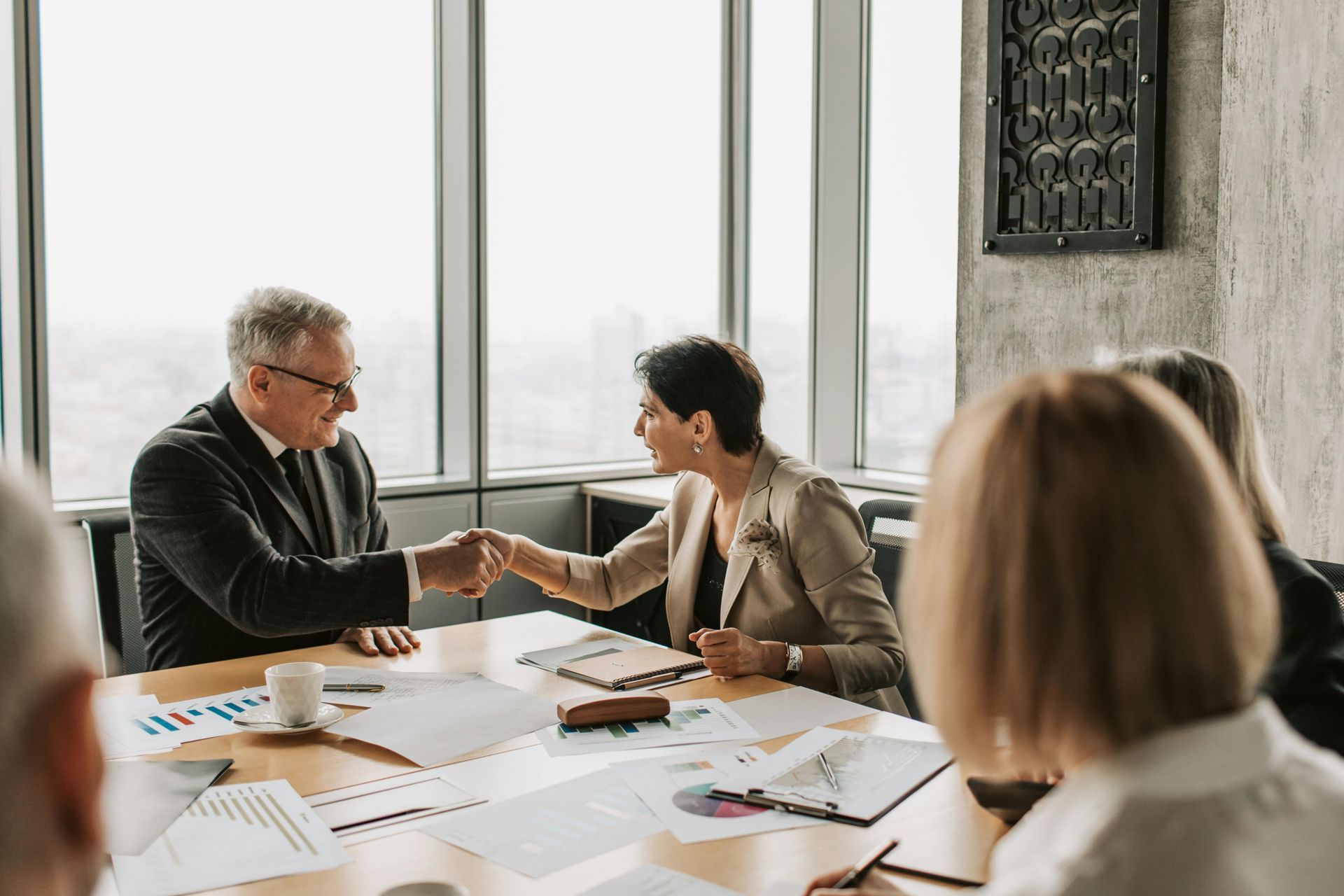 Two professionals in formal business attire shake hands across a conference table filled with documents in a bright office.