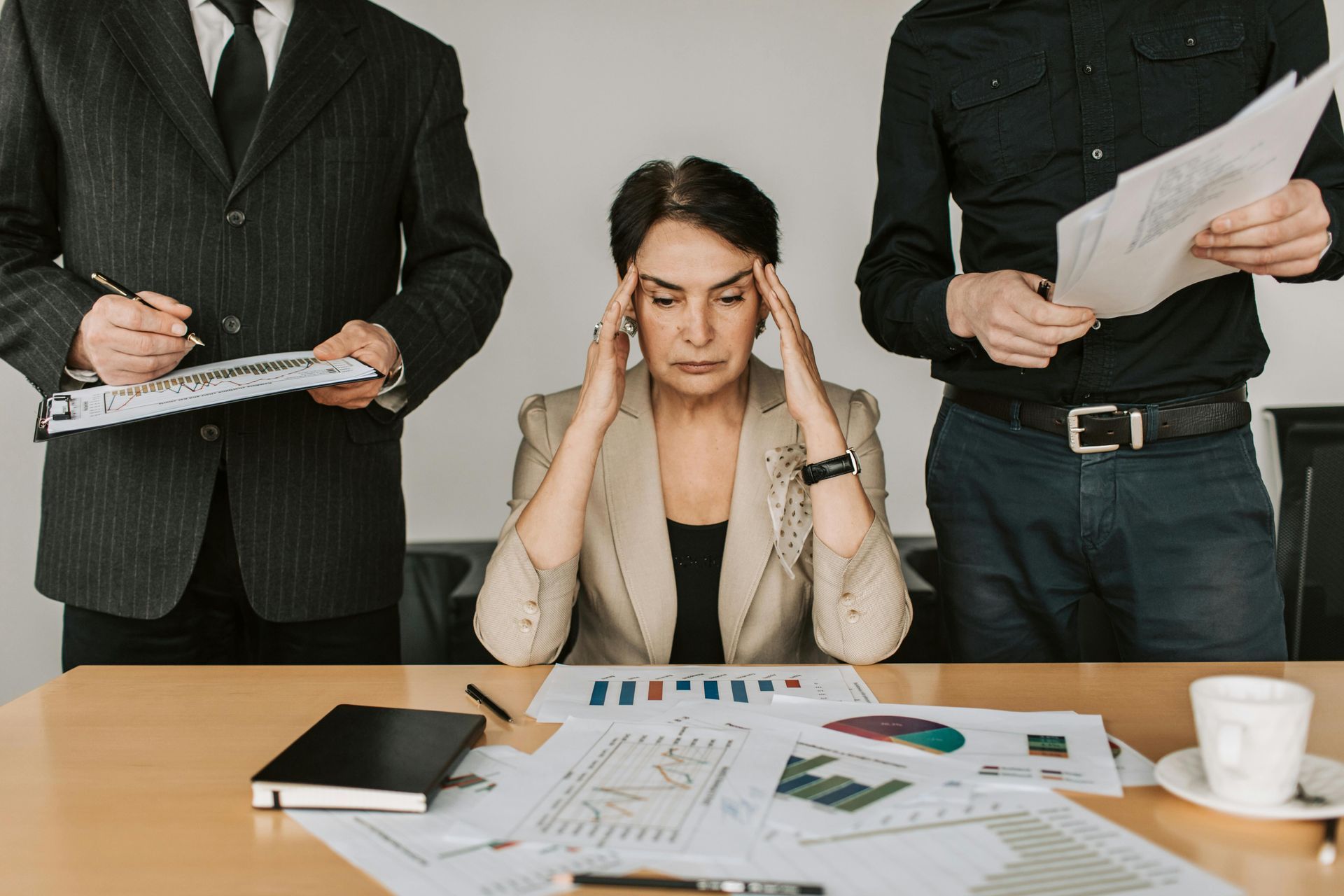 A stressed person holds their head at a desk covered in charts, flanked by two people holding documents and taking notes.