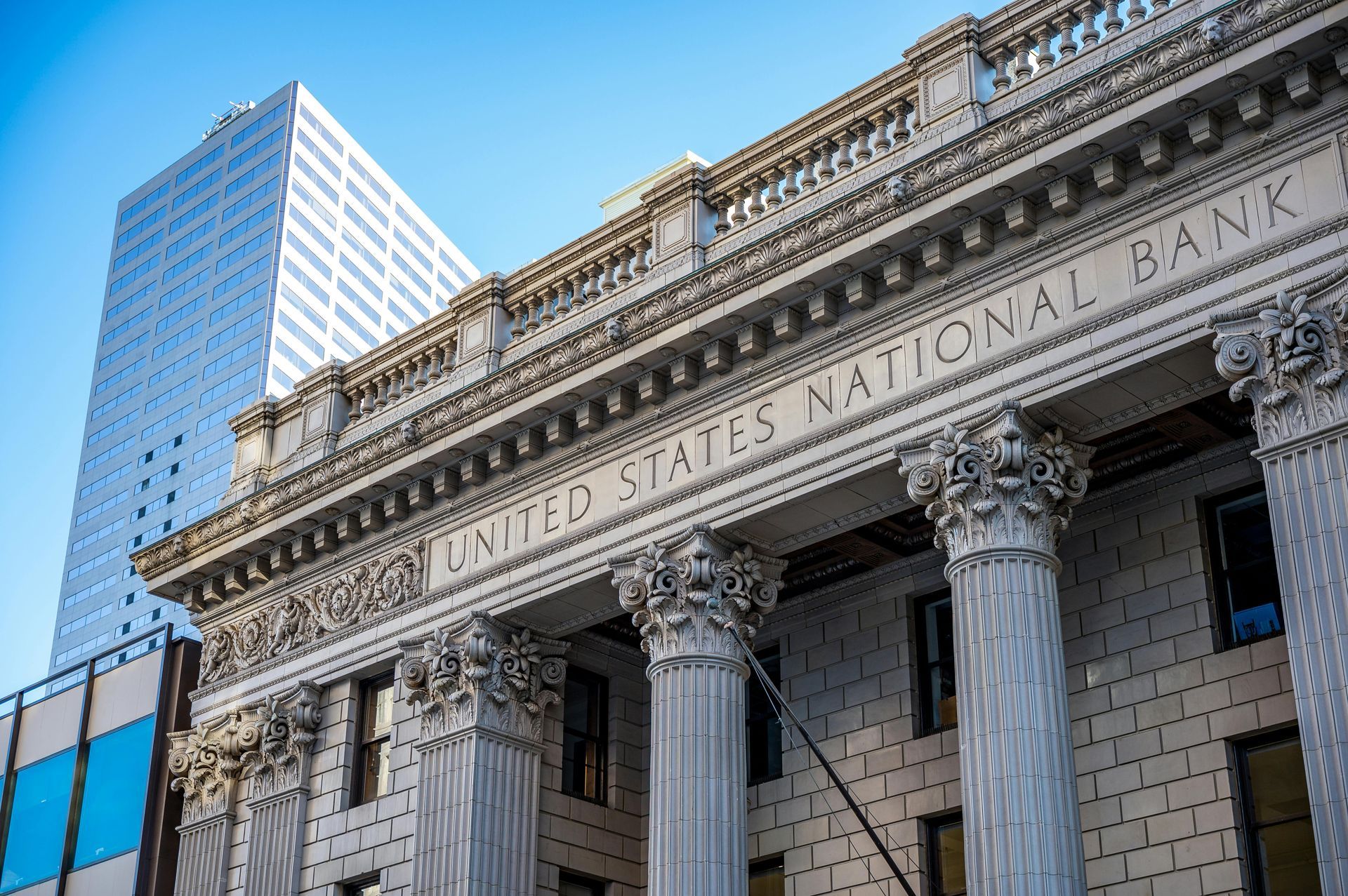 A low-angle view of the ornate U.S. National Bank building, with a modern glass skyscraper rising behind it.