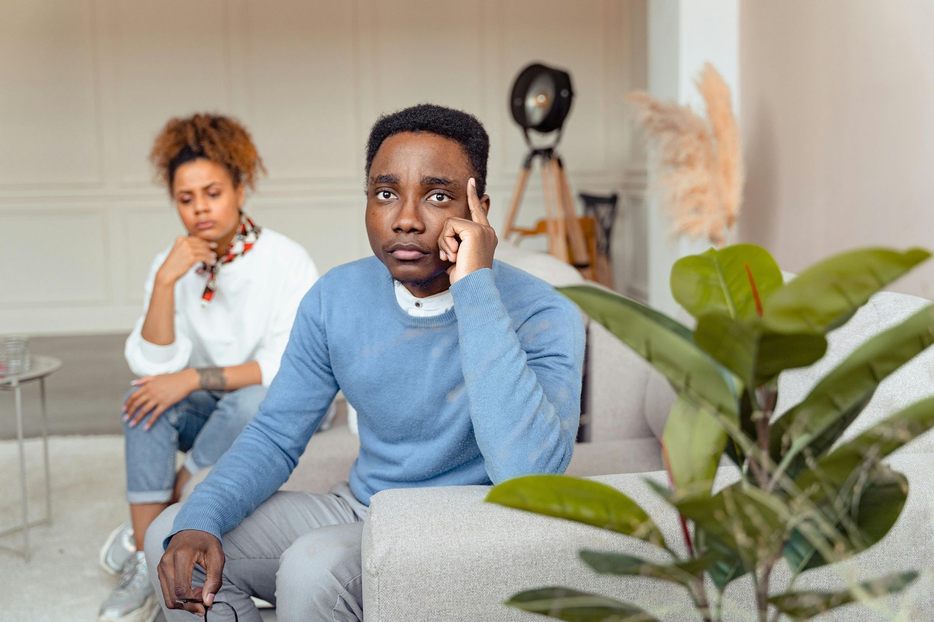Two people sit on a couch in a living room, looking thoughtful or concerned with their hands near their faces.