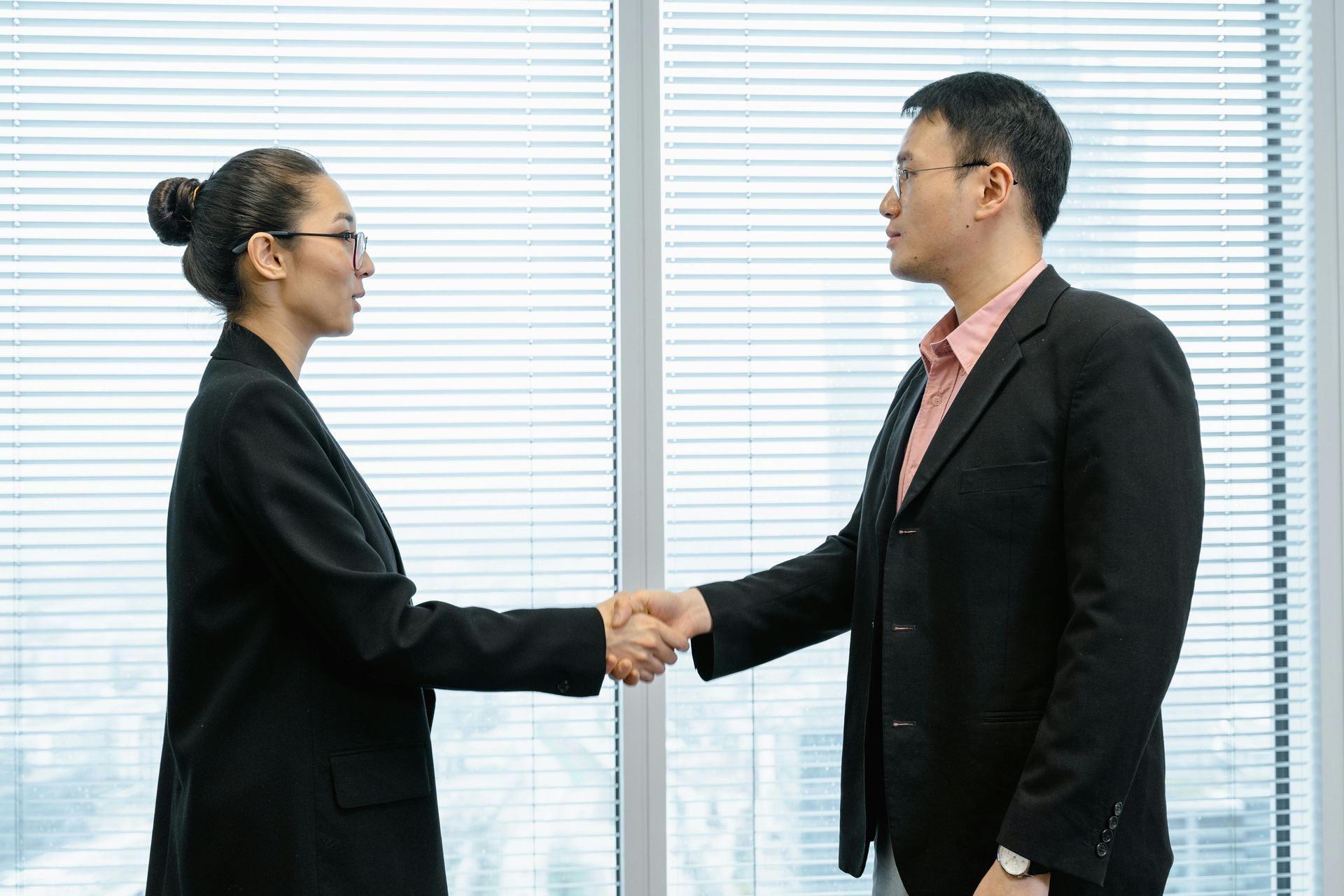 Two people in business suits shaking hands by a window.