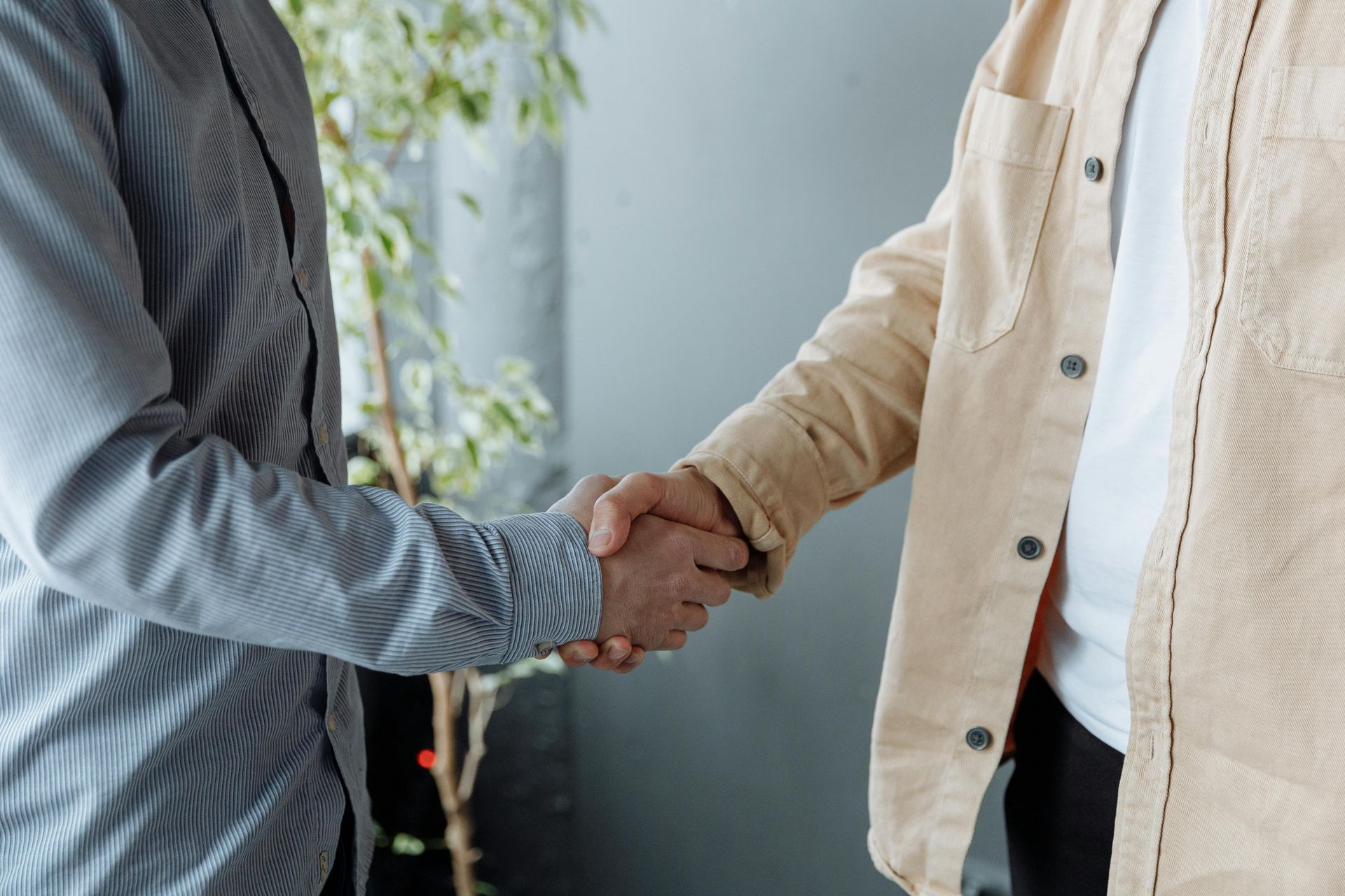 Two people shake hands, wearing casual button-down shirts, set against a grey wall with a small plant in the background.