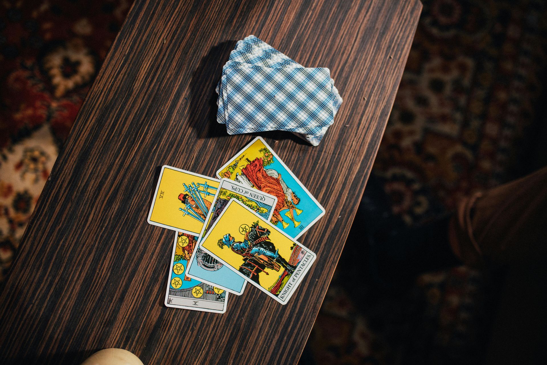 Tarot cards fanned out on a dark wood table with a deck of cards and a patterned rug in the background.