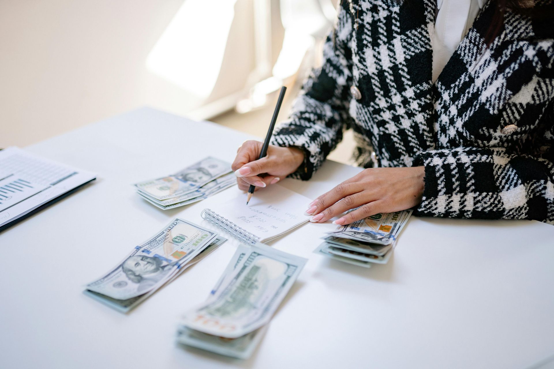 A person in a black and white plaid jacket writes on a notepad surrounded by stacks of US dollar bills on a white desk.