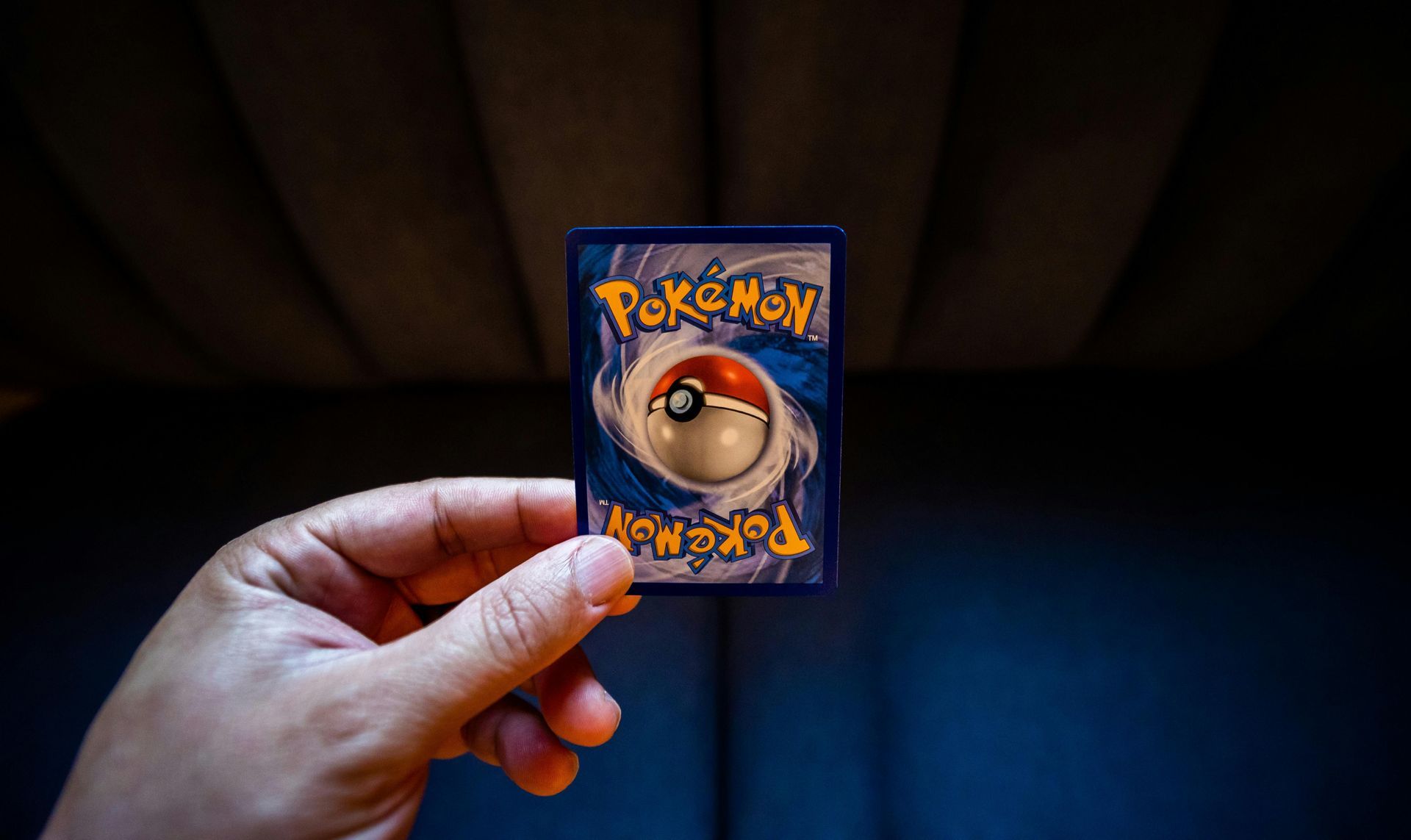 Hand holding a Pokémon card, showing the blue back with a Poké Ball design against a dark background.