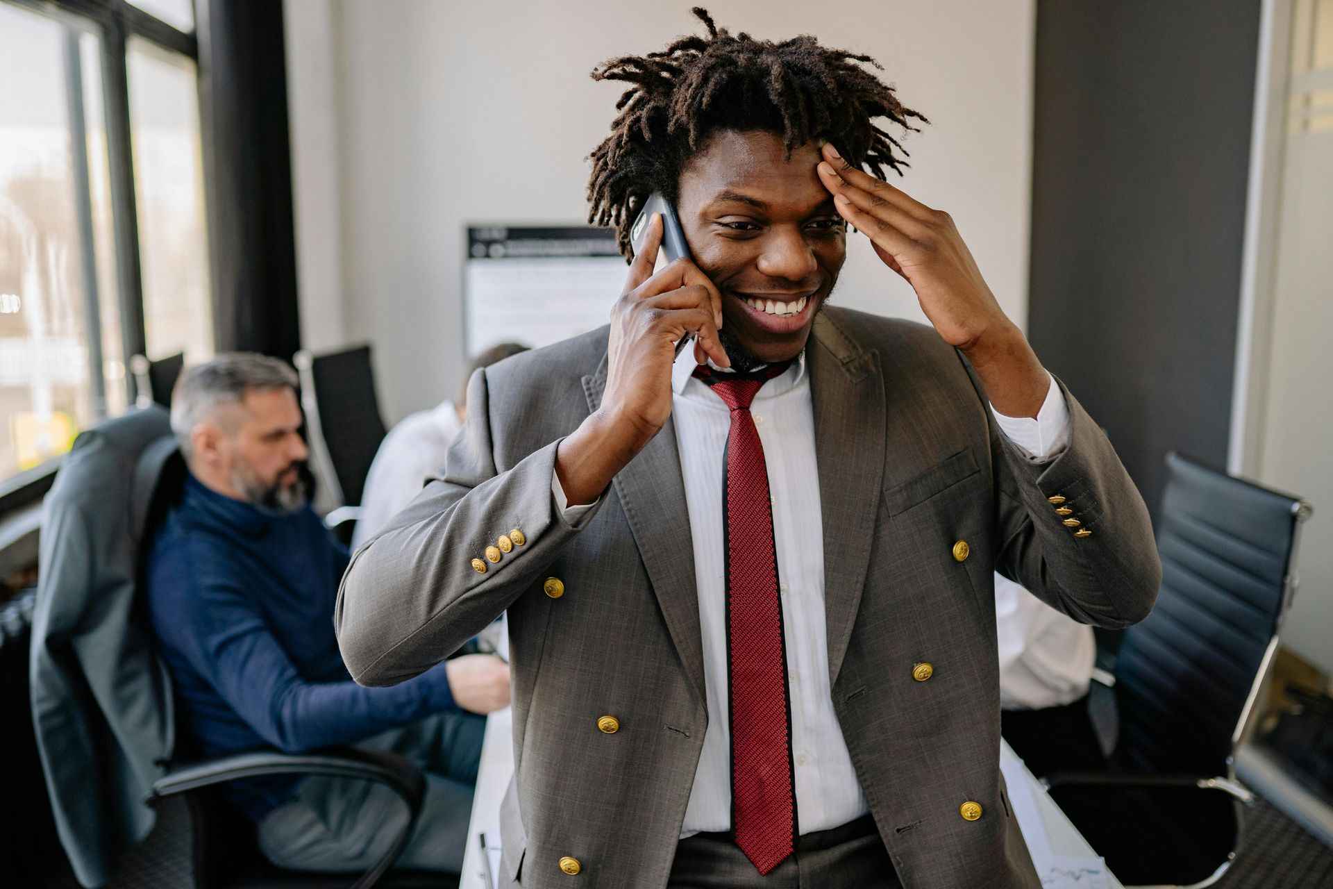 A person in a suit and tie talks on a phone in an office, smiling with one hand held to their forehead.