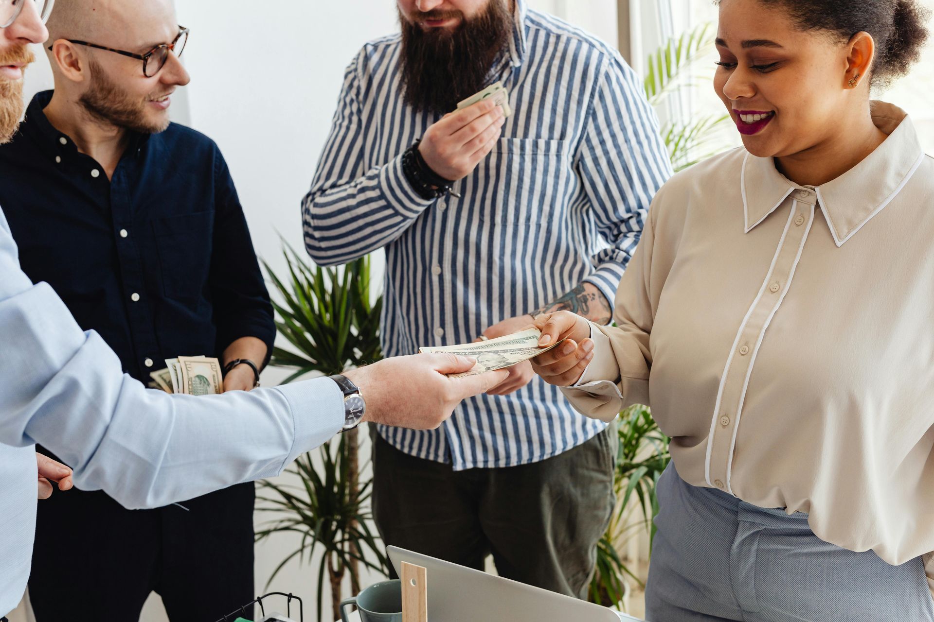 A person in a light blue shirt hands cash to a woman in a cream blouse while two others look on in an office setting.