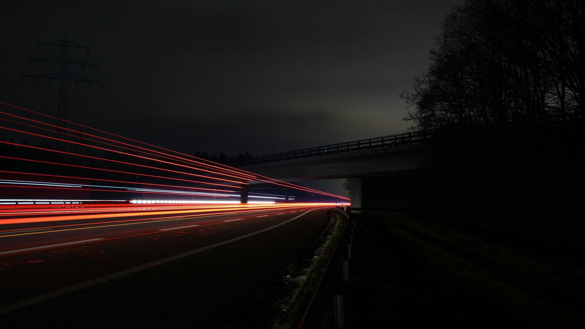 Long exposure of red taillights streaming along a dark highway passing under an overpass at night.