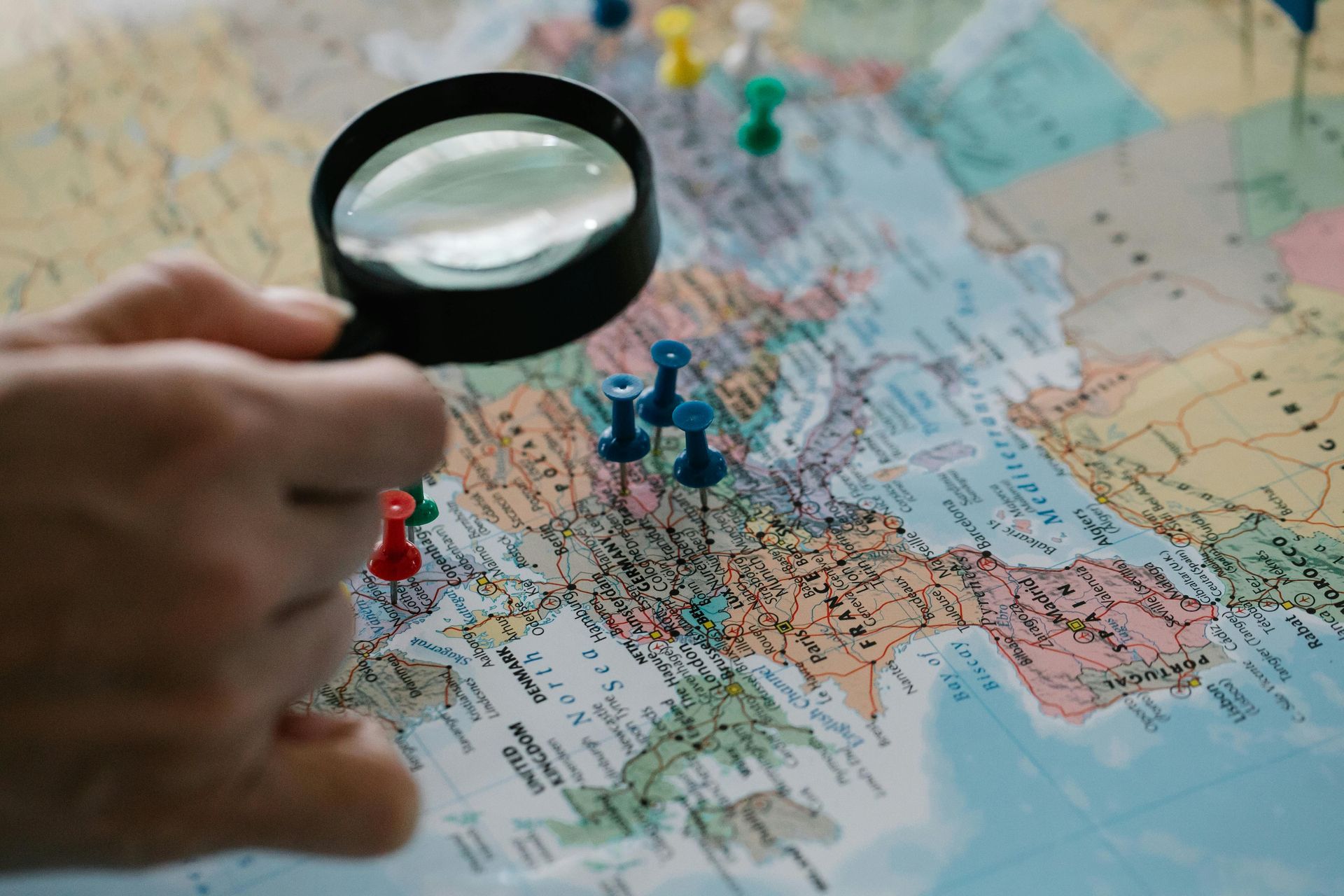 Hand holding magnifying glass over a map with push pins marking locations.