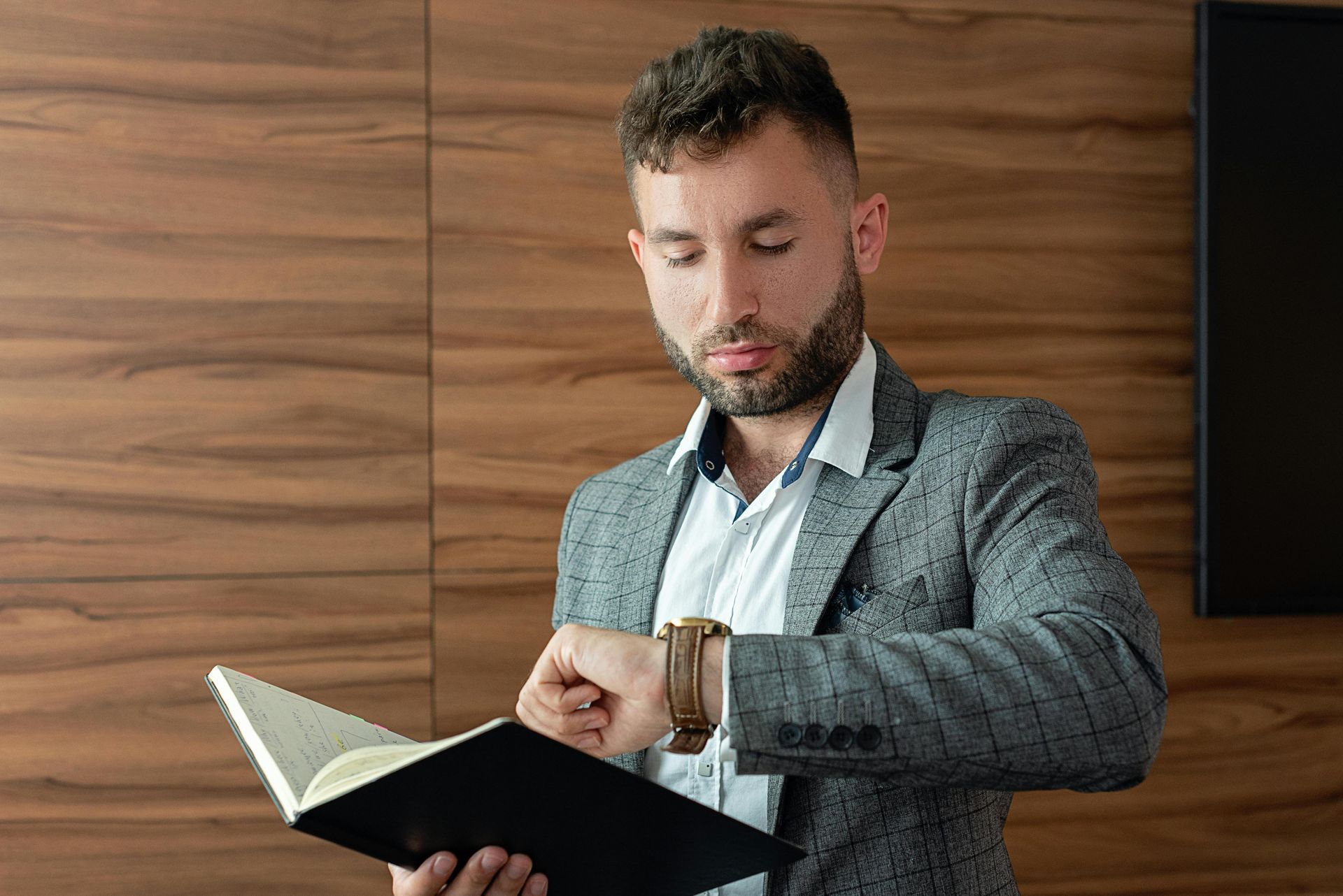 Man in suit jacket looking at his watch, holding a notebook, wood-paneled background.