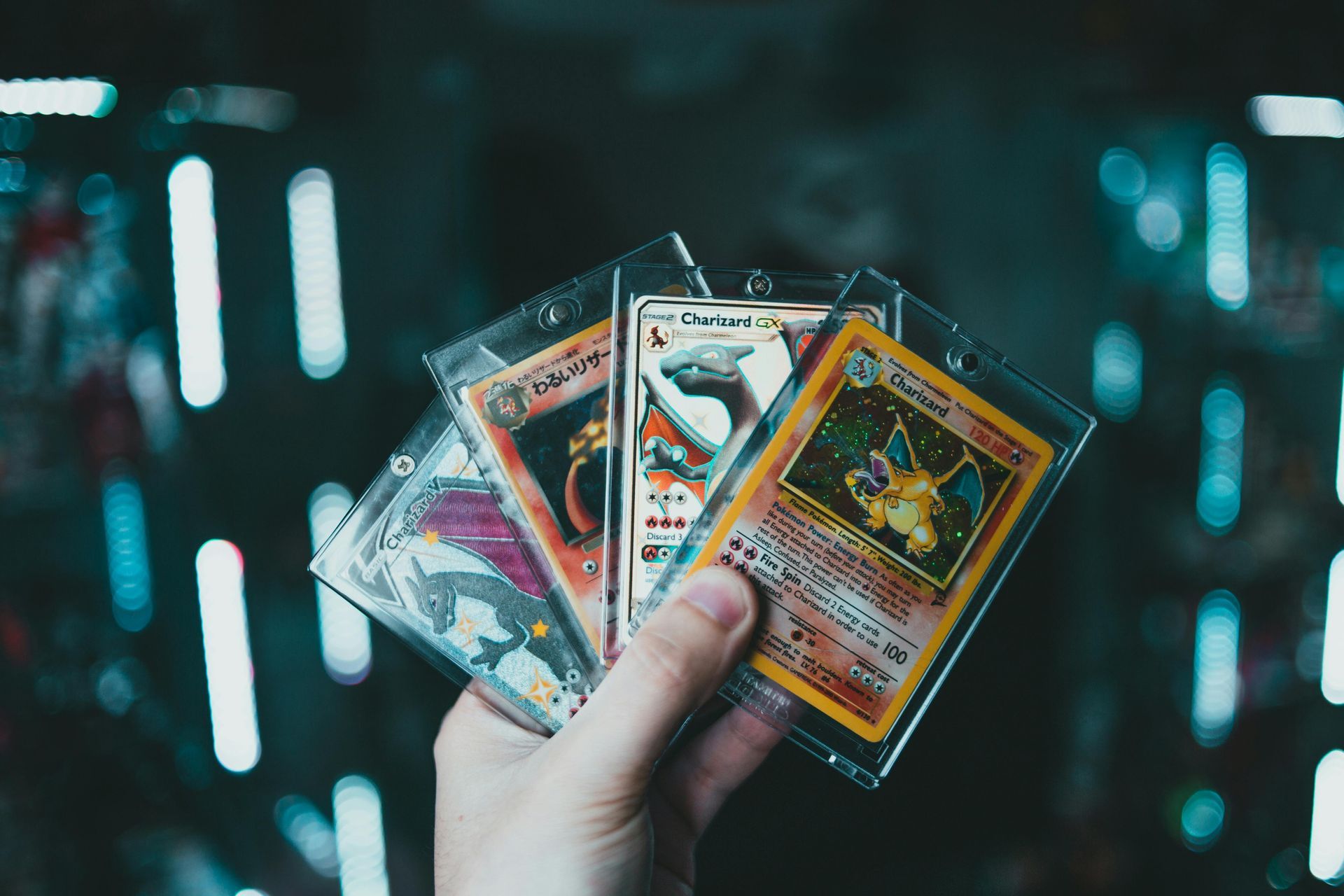 Hand holding four Pokémon cards in protective sleeves, against a blurry, lit-up background.