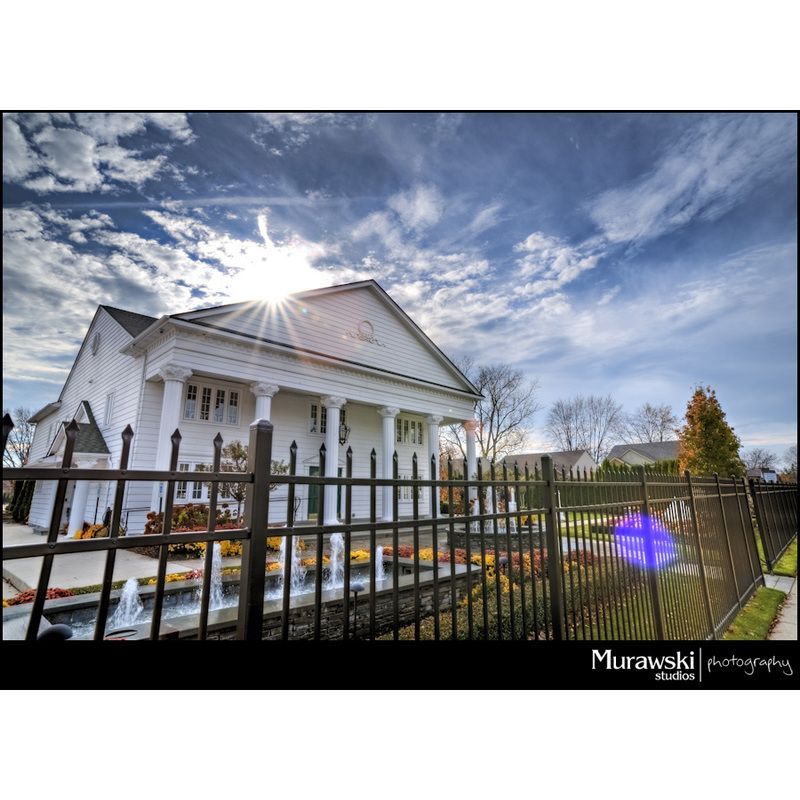 A large white house behind a fence with the sun shining through the clouds