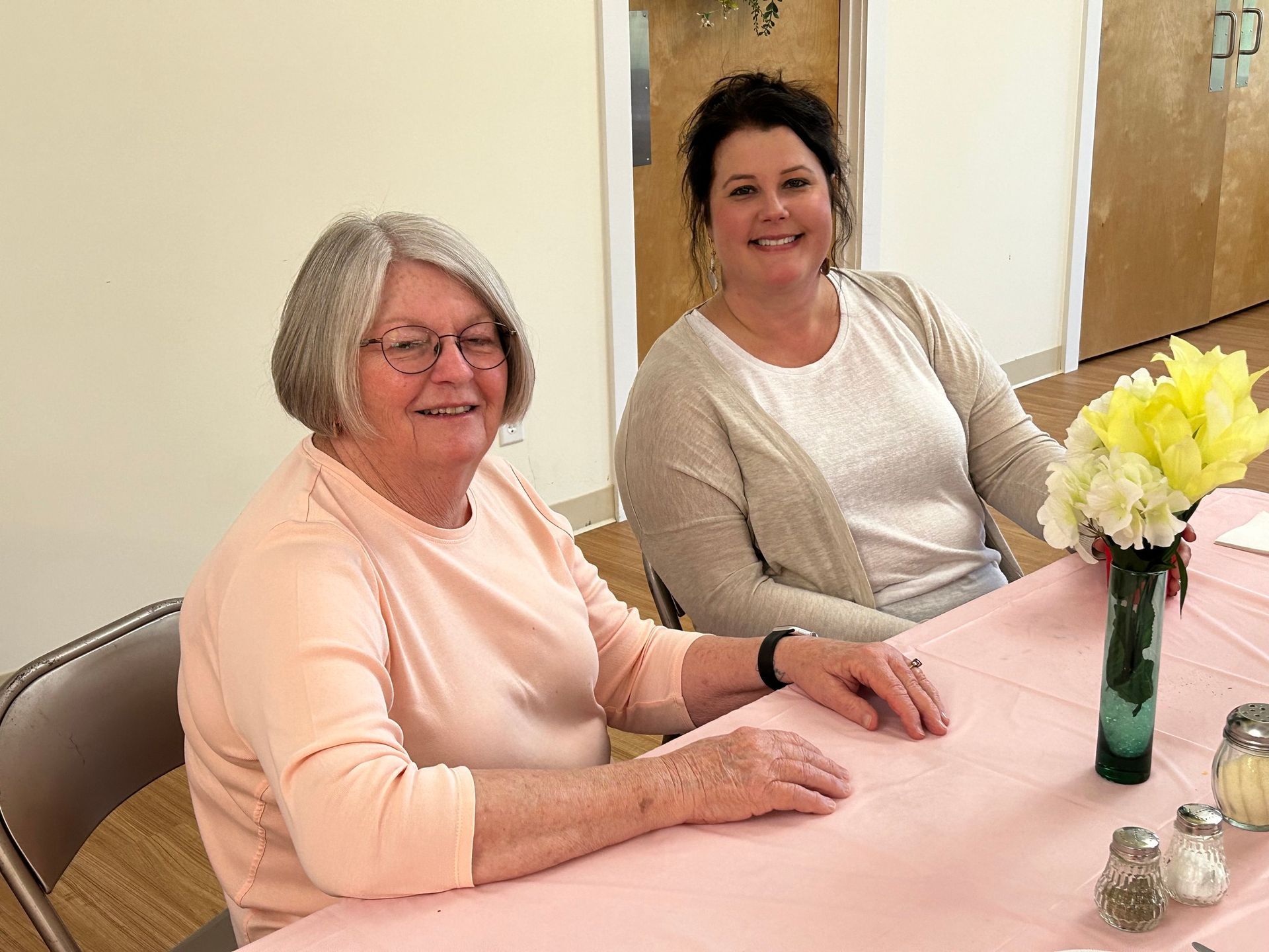 Two women are sitting at a table with flowers in a vase.