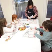 A group of children are sitting at a table with a woman standing behind them.