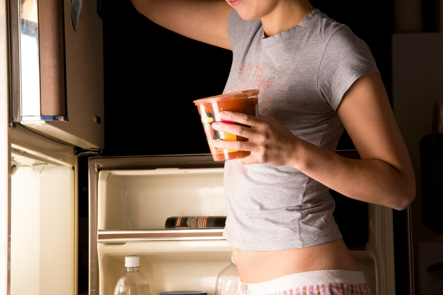 A woman is standing in front of an open refrigerator holding a cup of food.