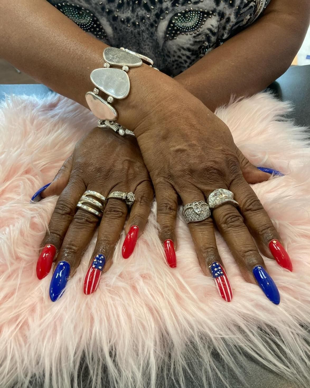 Hands with red, white, and blue American flag nail art, silver jewelry on pink fur.