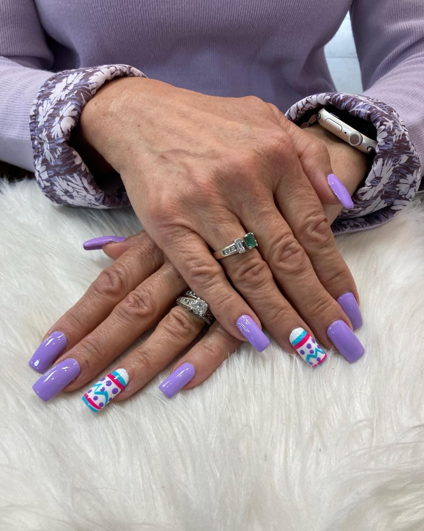 Hands with lavender-colored square nails, two decorated with colorful winter sweater patterns.