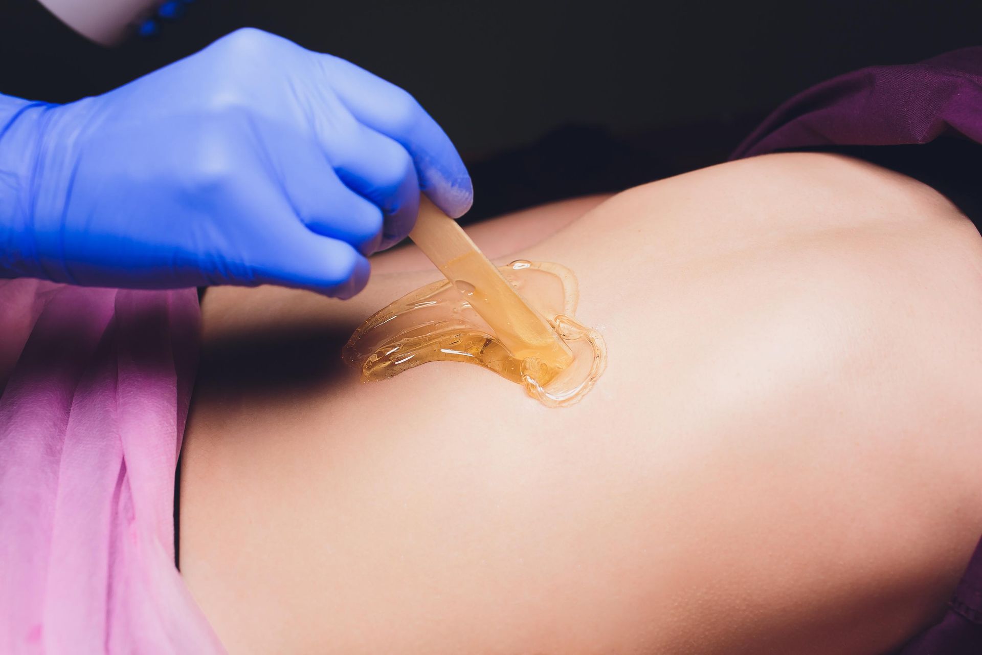 A gloved hand applies wax to a person's skin with a wooden stick in a salon setting.