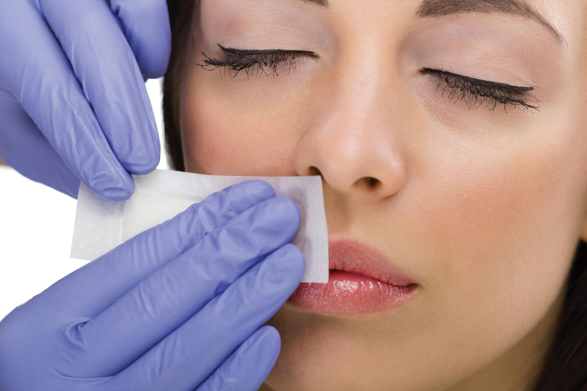 Hands in blue gloves holding a waxing strip to a woman's upper lip.