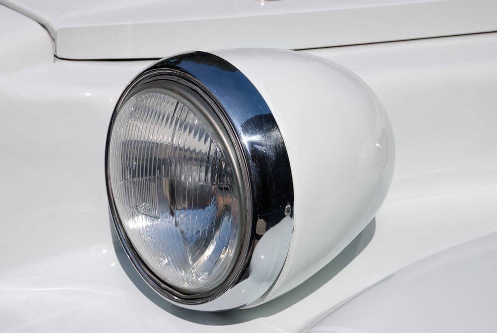 Front detail of a classic round headlight with chrome surround on a polished white exotic car.
