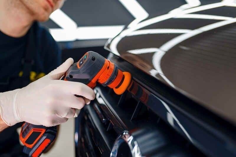Professional detailer in white gloves polishes a dark car's grille with a compact orbital buffer.
