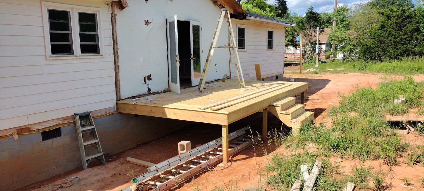 A house with a wooden deck under construction, reddish-brown dirt, and grass.