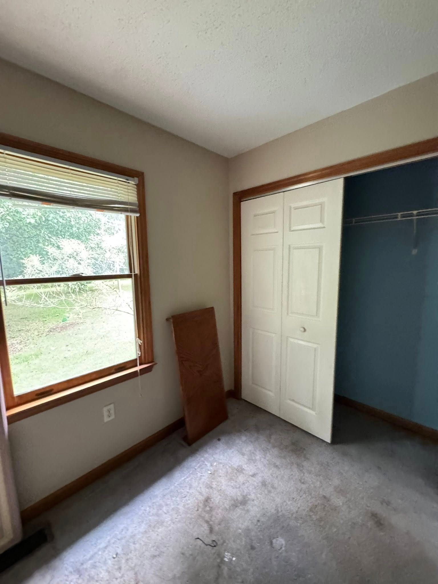 Empty bedroom with window, closet, tan walls, and gray carpet.