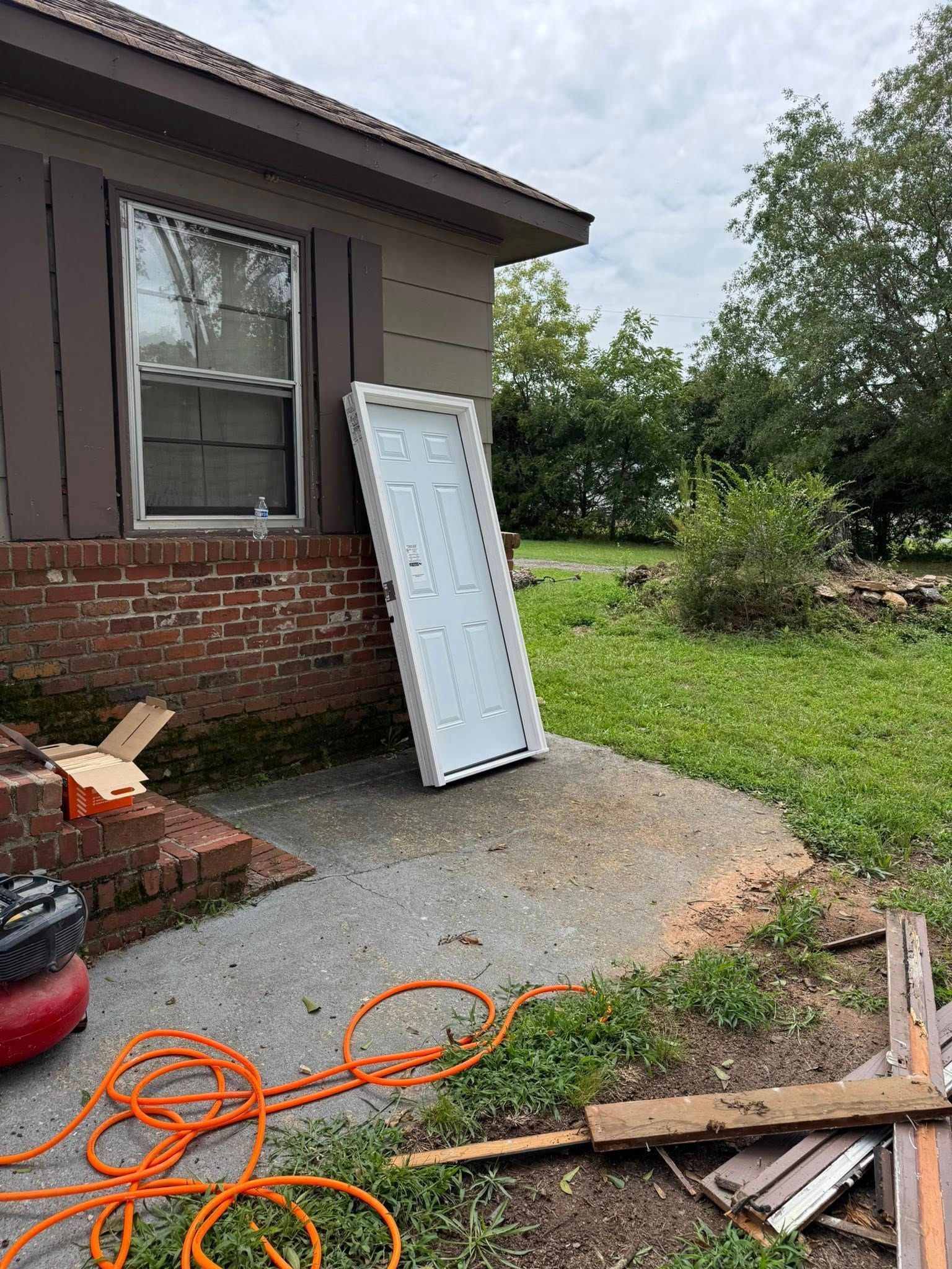 White door propped against a brown building, next to a window and concrete slab. Tools and yard debris in view.