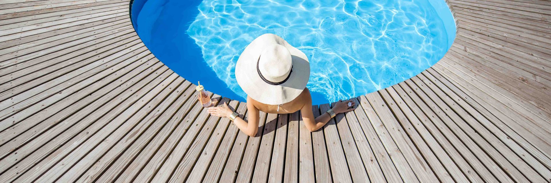 A woman chilling in her circular pool with a sun hat on and a drink by her hand on the deck