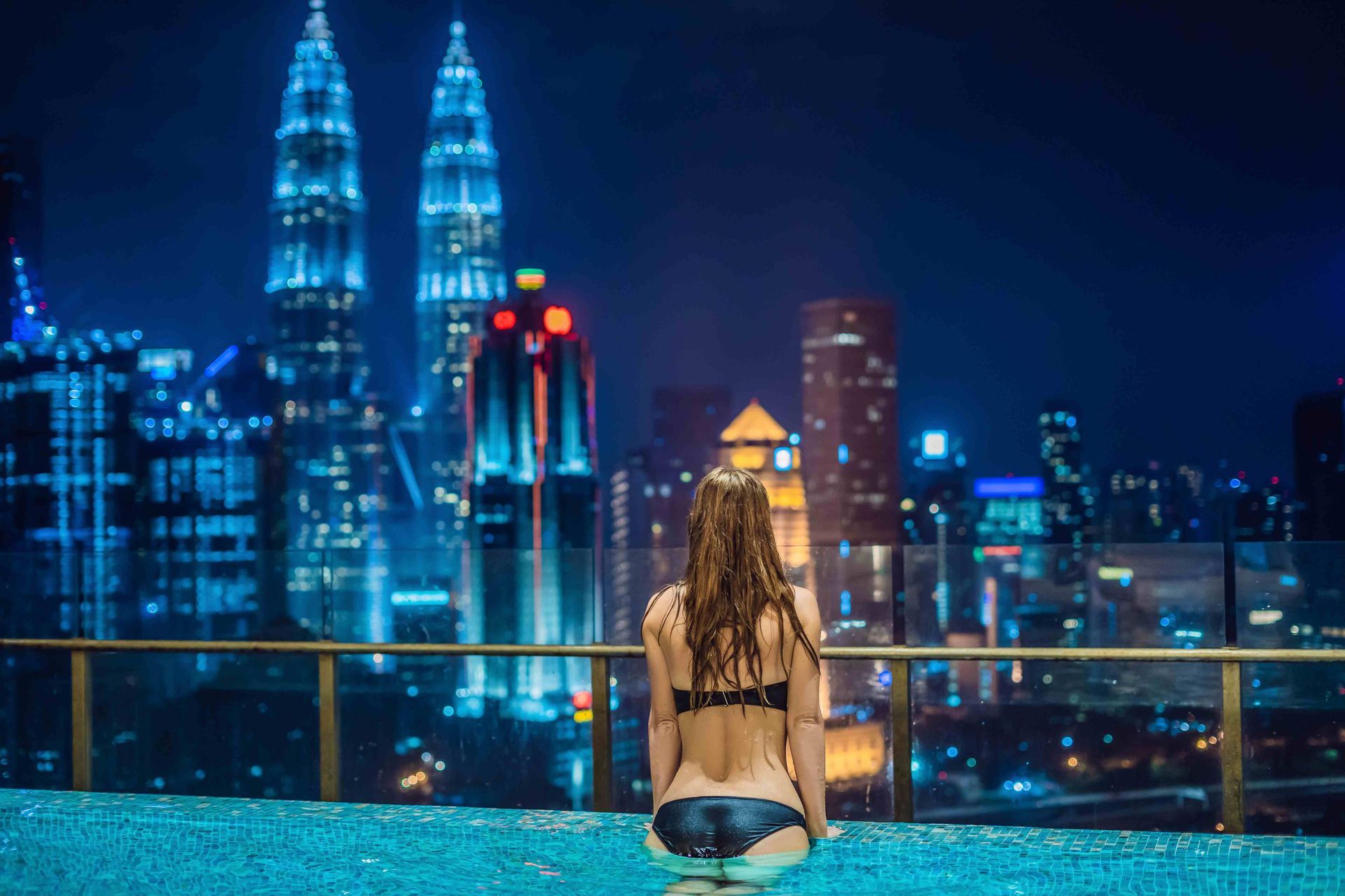 A pretty woman standing in an infinity pool facing the city