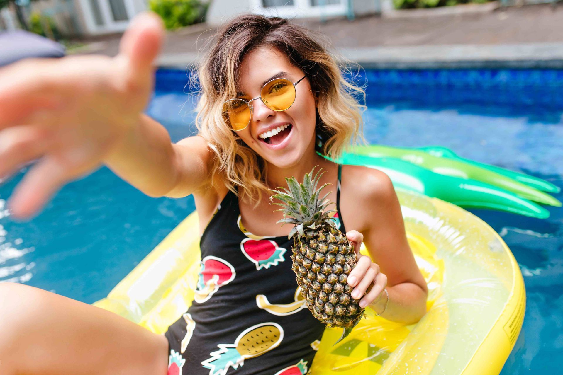 A woman on a raft in a swimming pool with a pineapple in her hand, reaching out toward the camera
