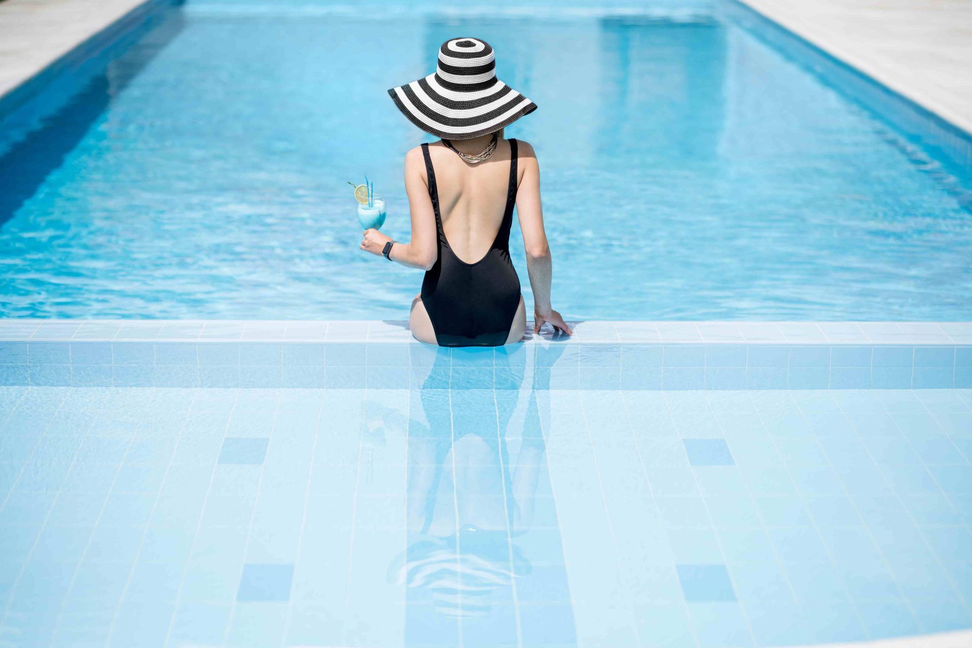 A woman sitting on the edge of a pool, wearing a striped sunhat