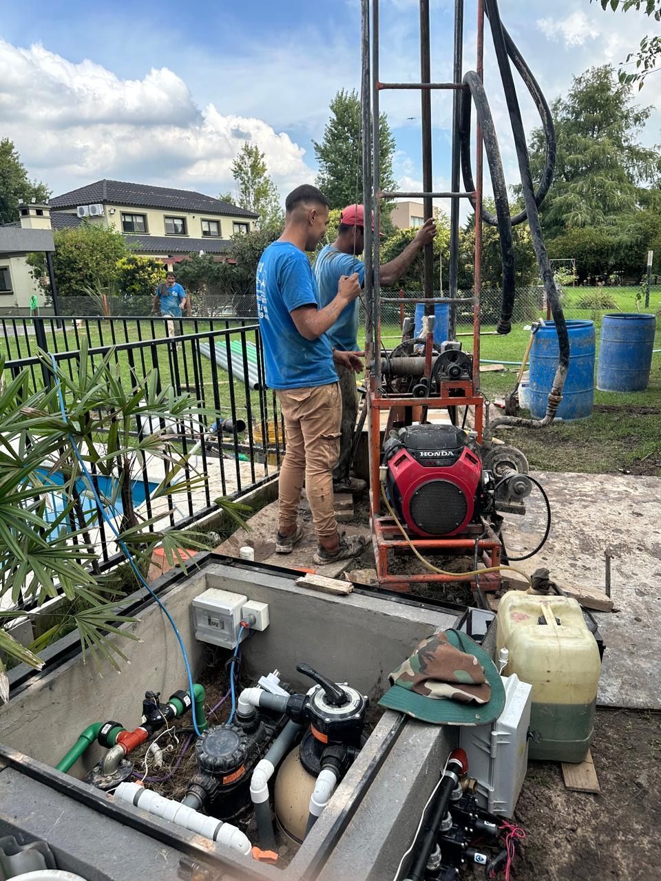 Dos hombres están trabajando en un pozo en un patio trasero al lado de una piscina.