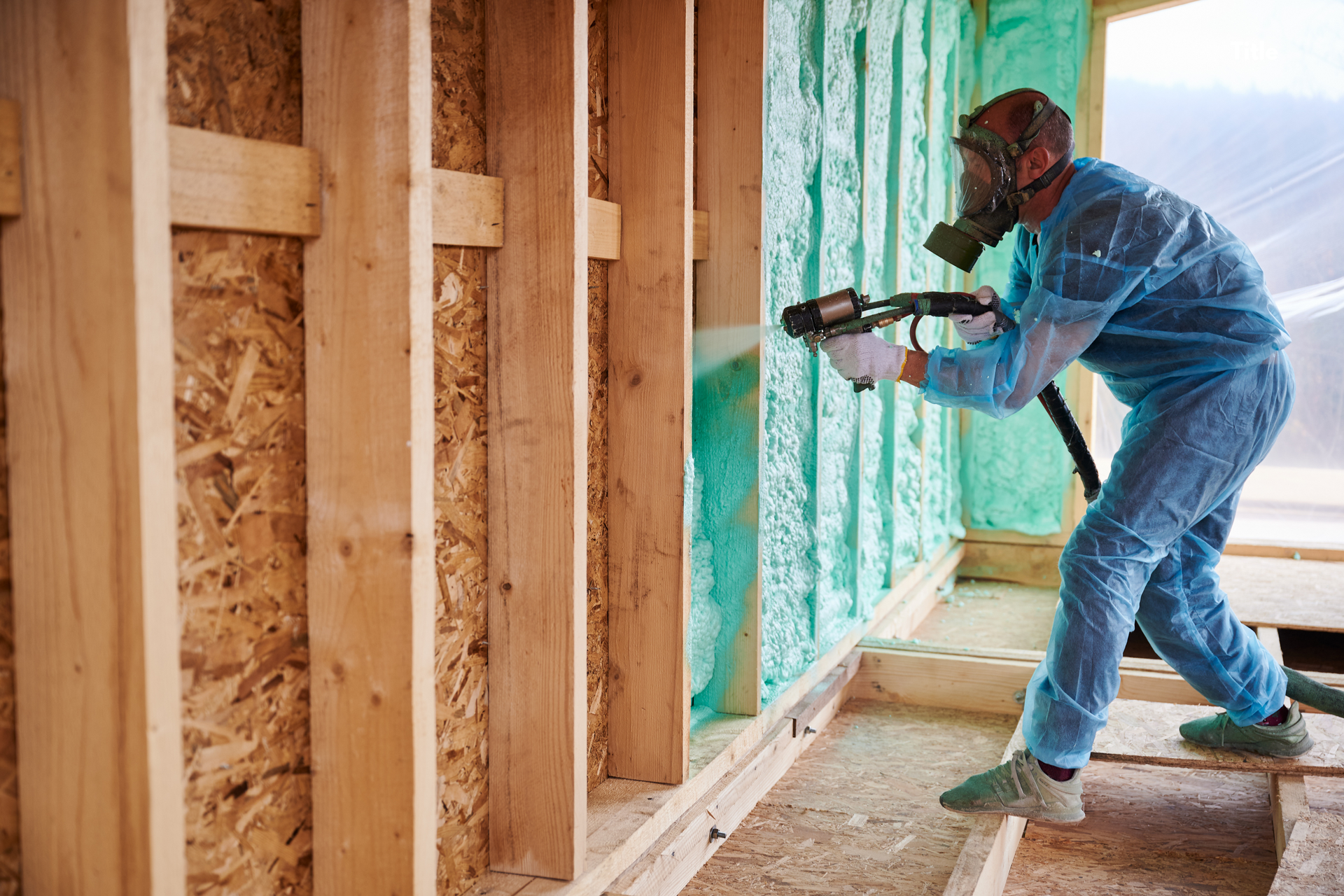 Person in protective suit spraying insulation in a wood-framed room.