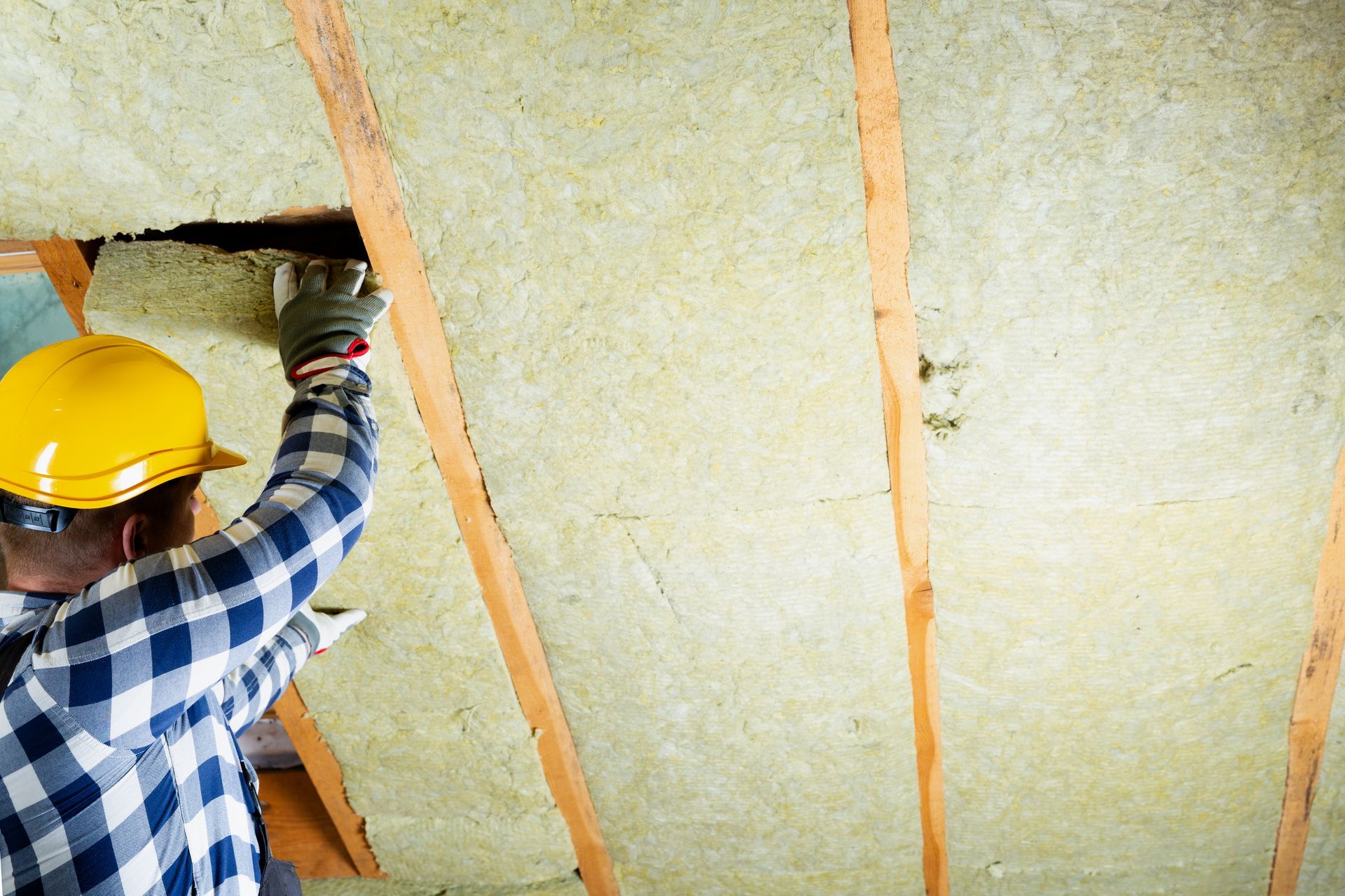 A construction worker in a yellow hard hat and plaid shirt installs yellow insulation between wooden ceiling rafters.