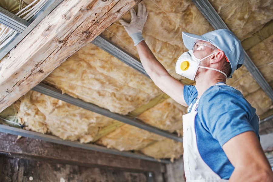 A worker with a mask and gloves installs insulation in a ceiling.
